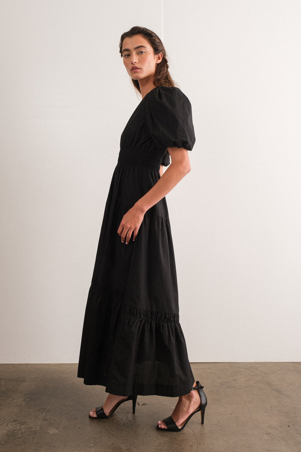 woman wearing a black puff sleeve maxi dress with a gathered waist and tiered hem, paired with black high-heeled sandals