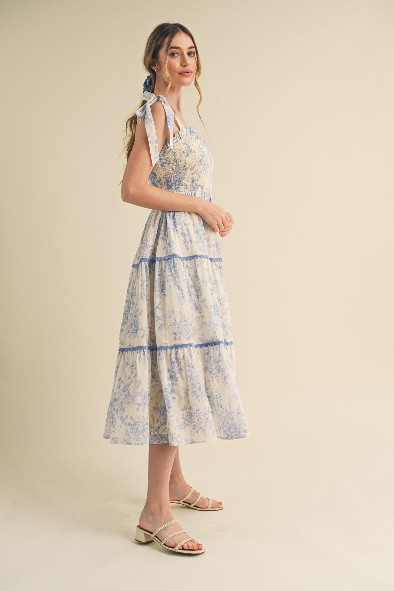 woman wearing a floral printed midi dress with blue accents and tie straps, standing sideways against a light background