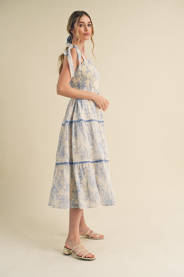woman wearing a floral printed midi dress with blue accents and tie straps, standing sideways against a light background