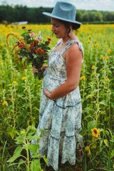 woman wearing a floral maternity maxi dress, holding a bouquet of flowers and cradling her baby bump in a sunflower field