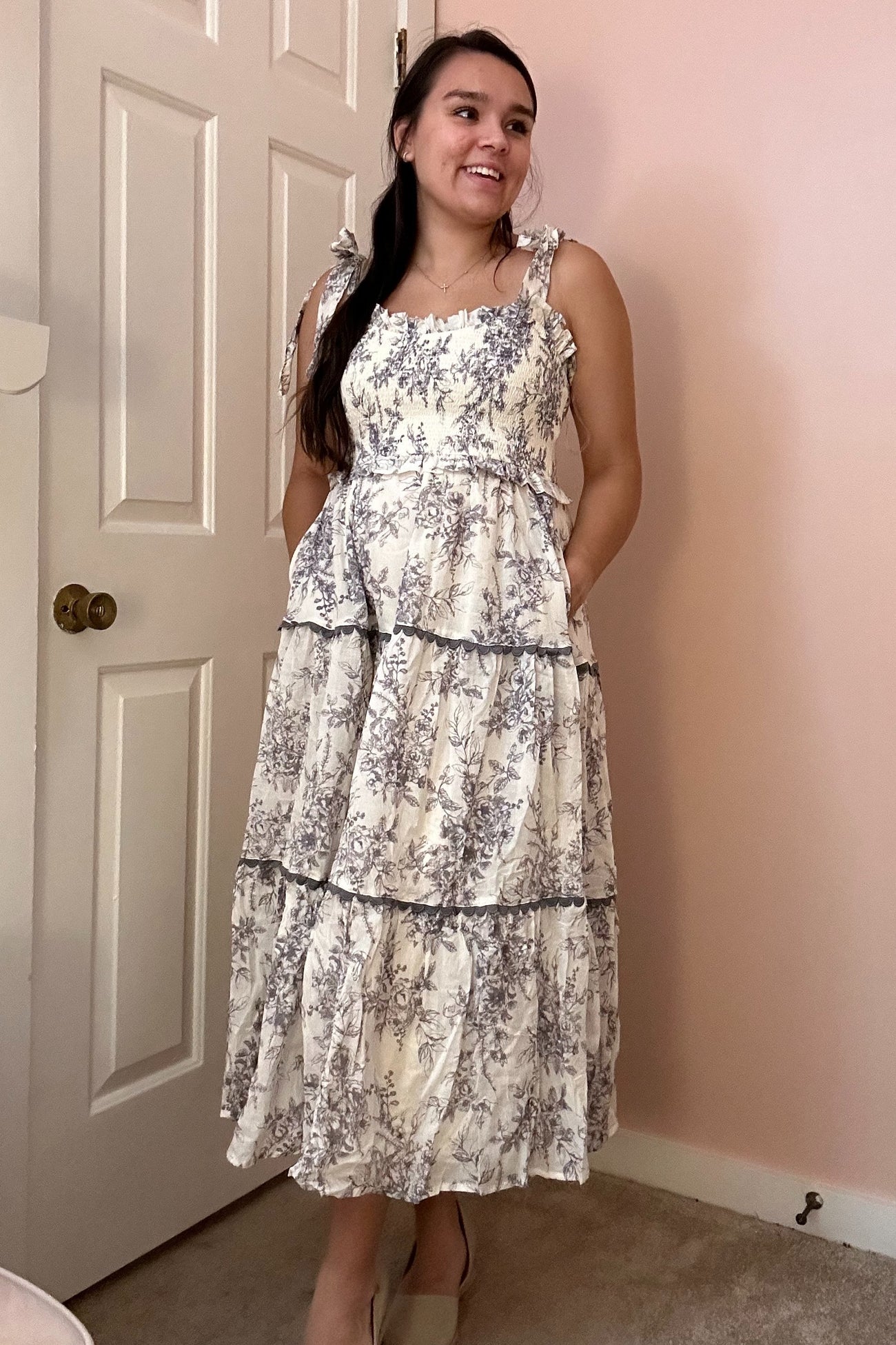 woman wearing a floral patterned midi dress with tiered layers and adjustable straps, standing against a pink wall
