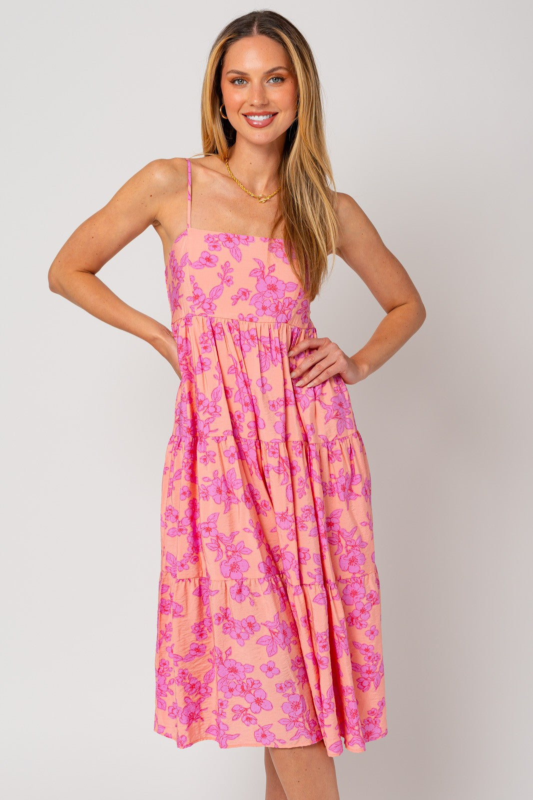 woman wearing a pink floral midi dress with thin straps and a tiered skirt, smiling with hands on her hips