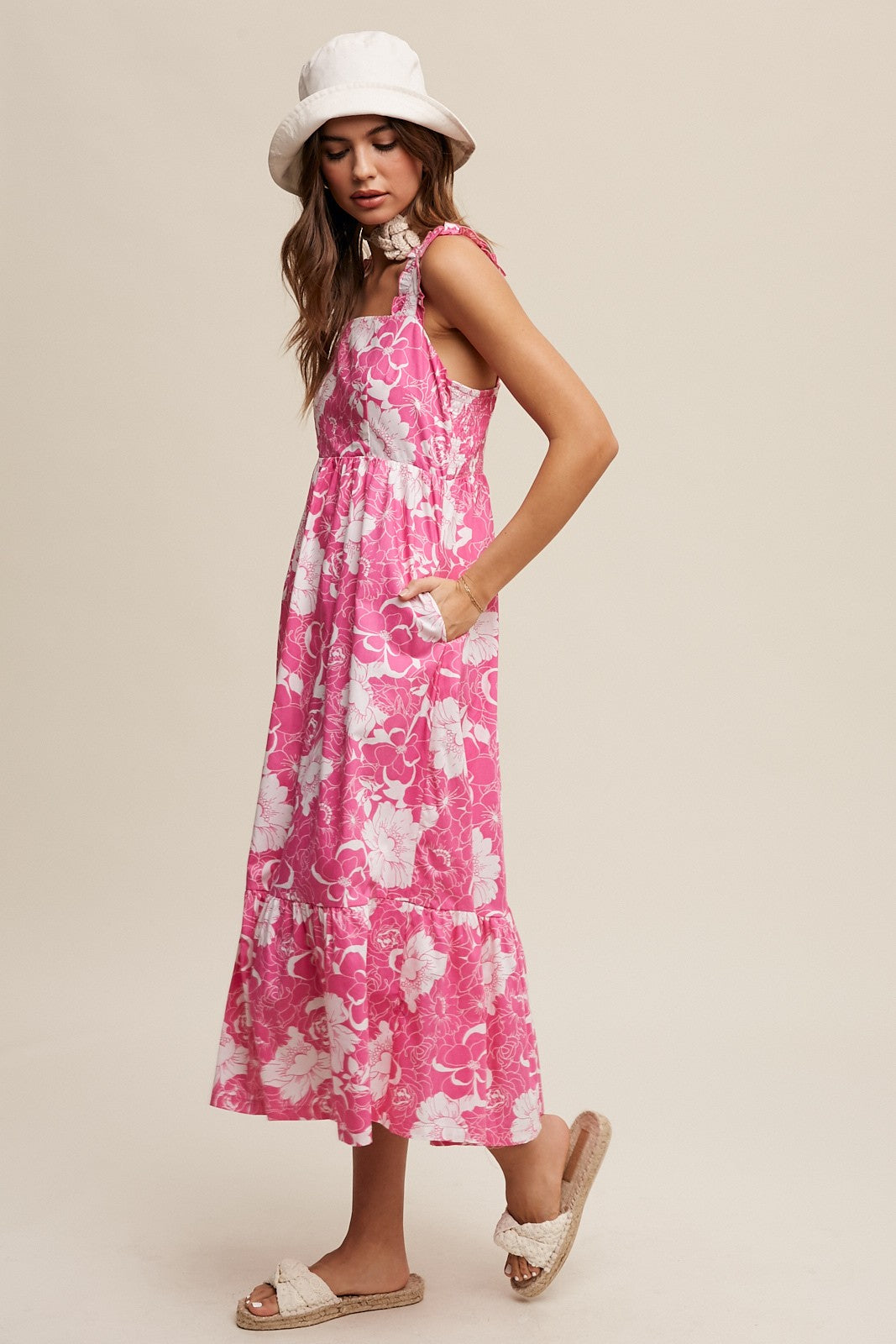 woman wearing a pink floral midi dress with pockets and a sun hat, paired with beige sandals