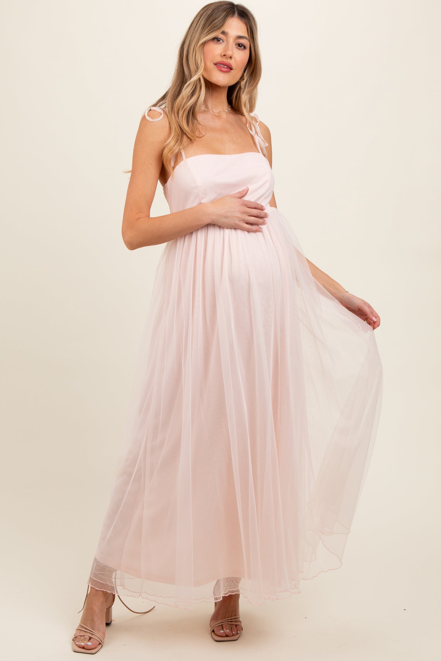 woman wearing a light pink maternity maxi dress with sheer overlay and adjustable straps, showing her baby bump
