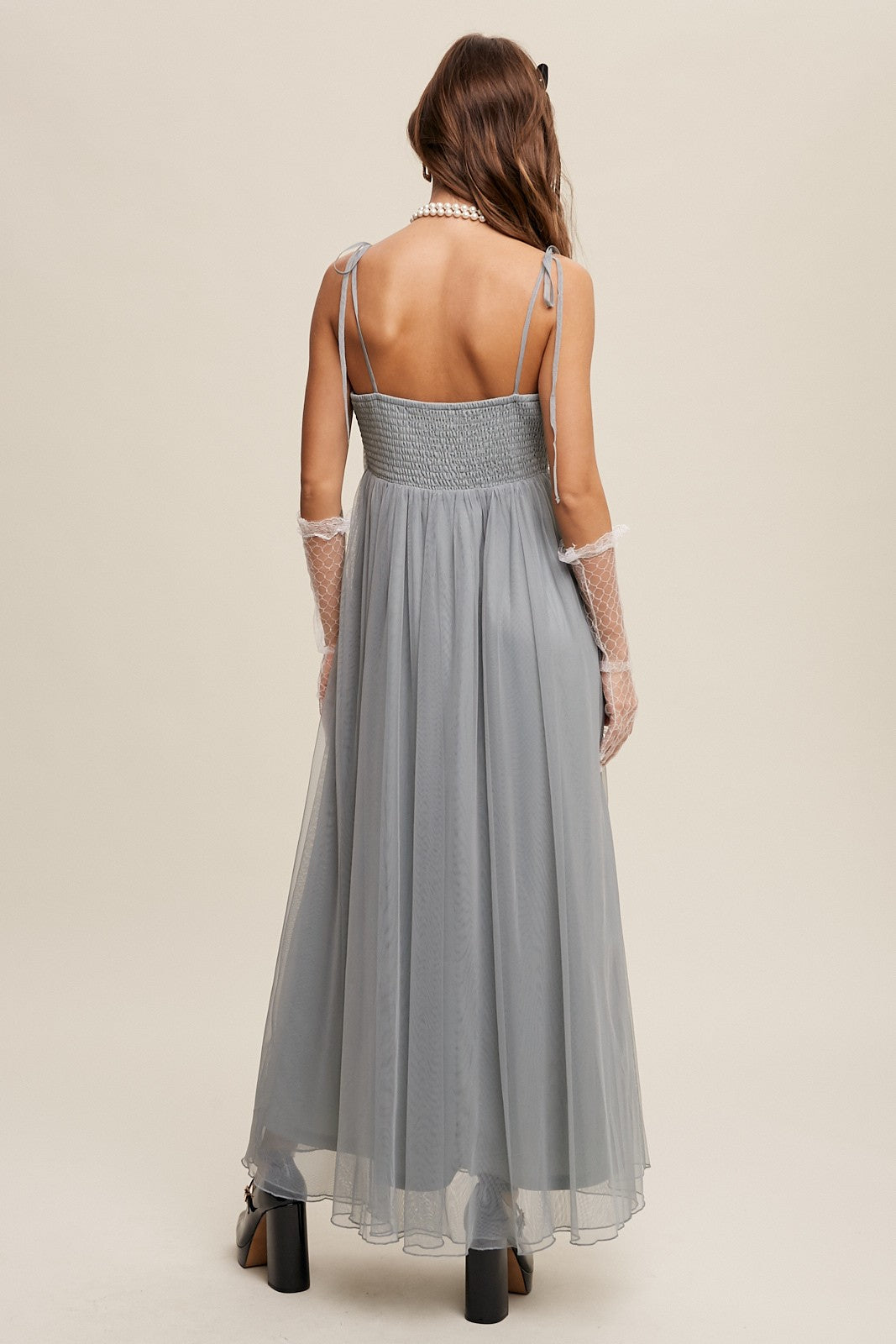 woman wearing a light grey long tulle dress with adjustable spaghetti straps and a smocked bodice, seen from the back
