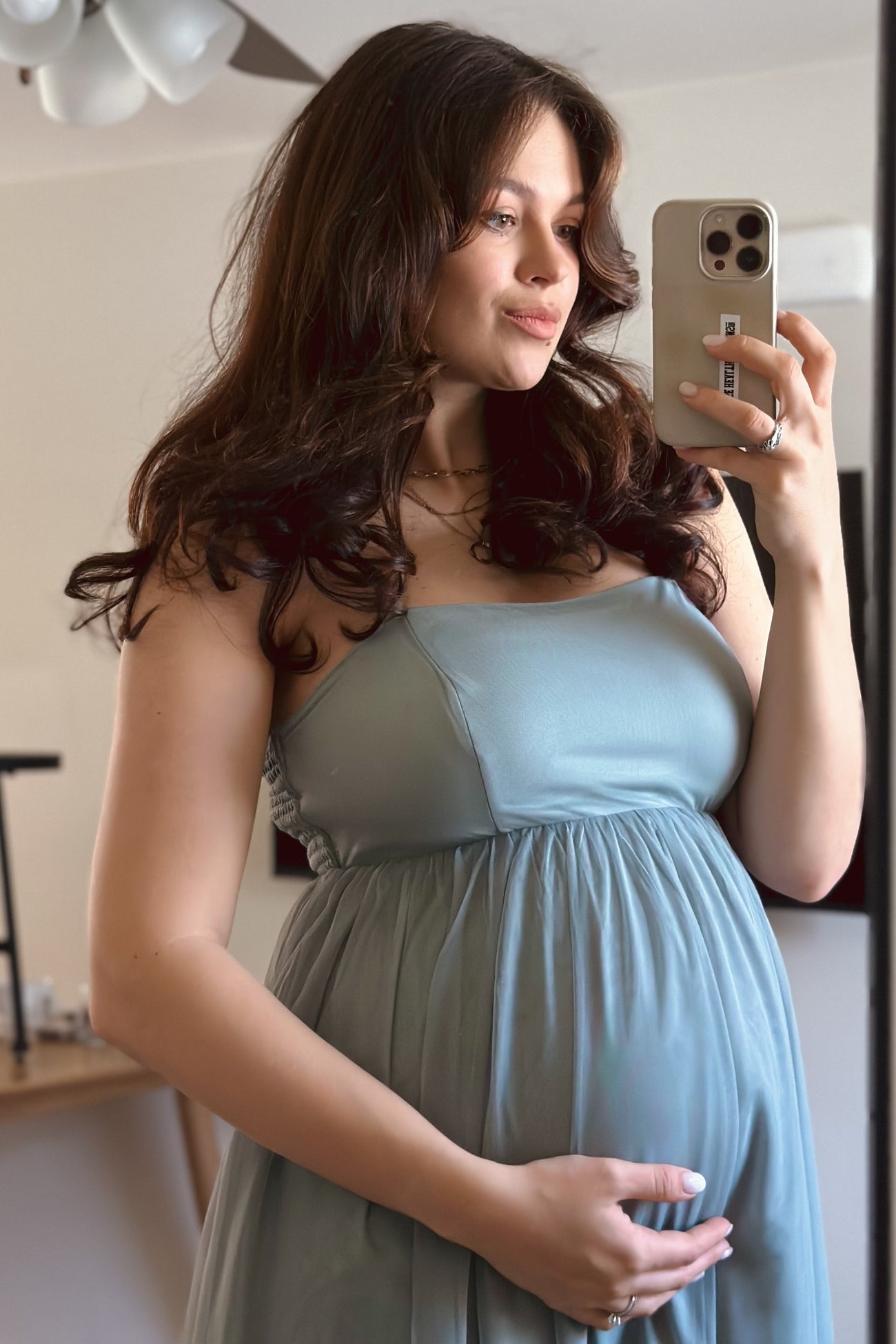 woman wearing a light blue maternity dress, cradling her baby bump and taking a selfie in front of a mirror