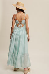 woman wearing a light green backless maxi dress with a tie detail, standing with a straw hat and platform sandals
