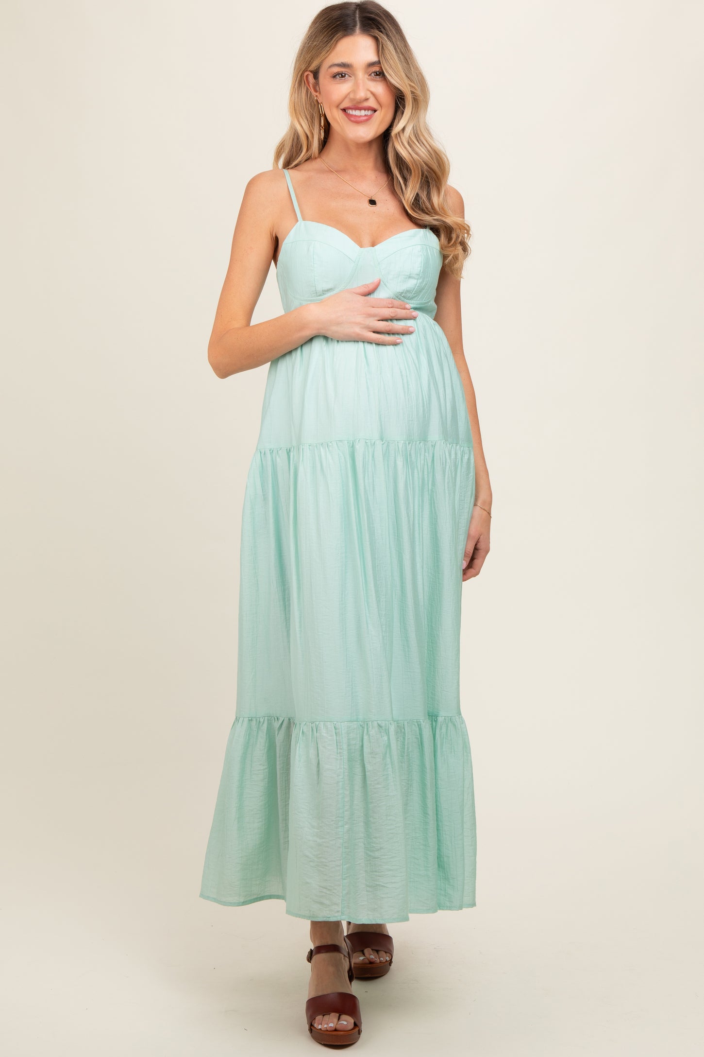 woman wearing a mint green maternity maxi dress with spaghetti straps and tiered detailing, holding her baby bump