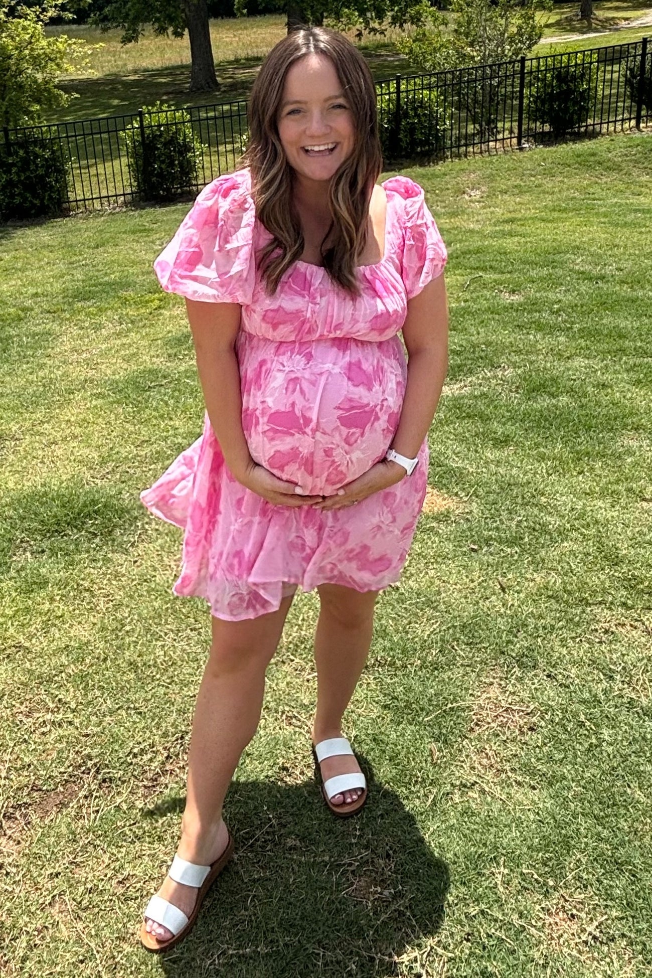 woman wearing a pink floral maternity dress and holding her baby bump outdoors on grass