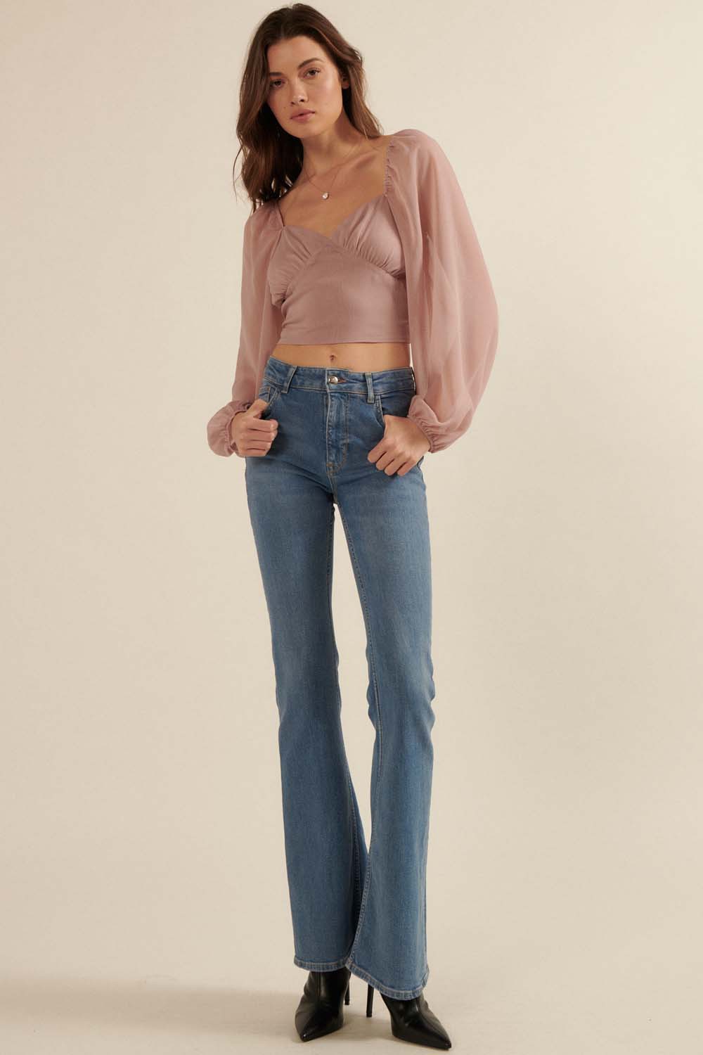 woman wearing a dusty rose cropped top with puff sleeves and high-waisted flared jeans, standing in heeled boots