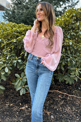 woman wearing a light pink long sleeve blouse with puffed sleeves and high-waisted blue jeans standing in front of greenery