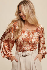 woman wearing a rust floral smocked top with long puff sleeves and a square neckline, paired with beige high-waisted trousers