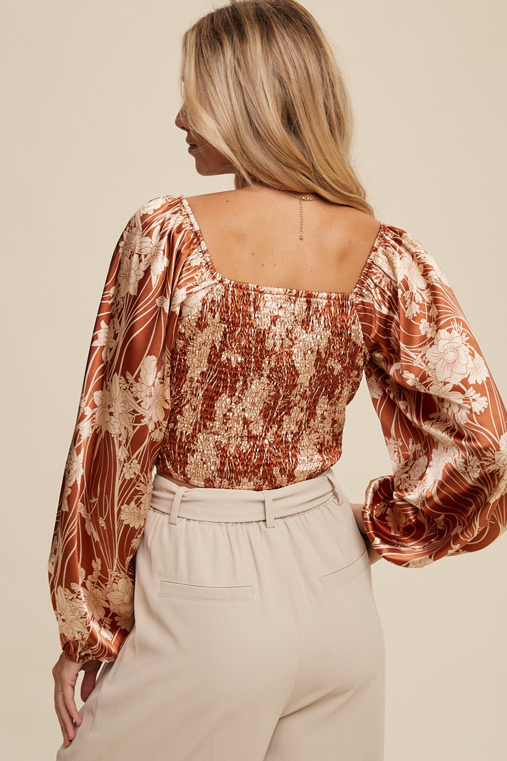 woman wearing a copper floral blouse with puff sleeves and smocked back paired with beige high-waisted trousers
