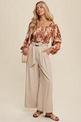 woman wearing a brown floral blouse with puff sleeves and beige high-waisted wide-leg trousers, holding a light bag