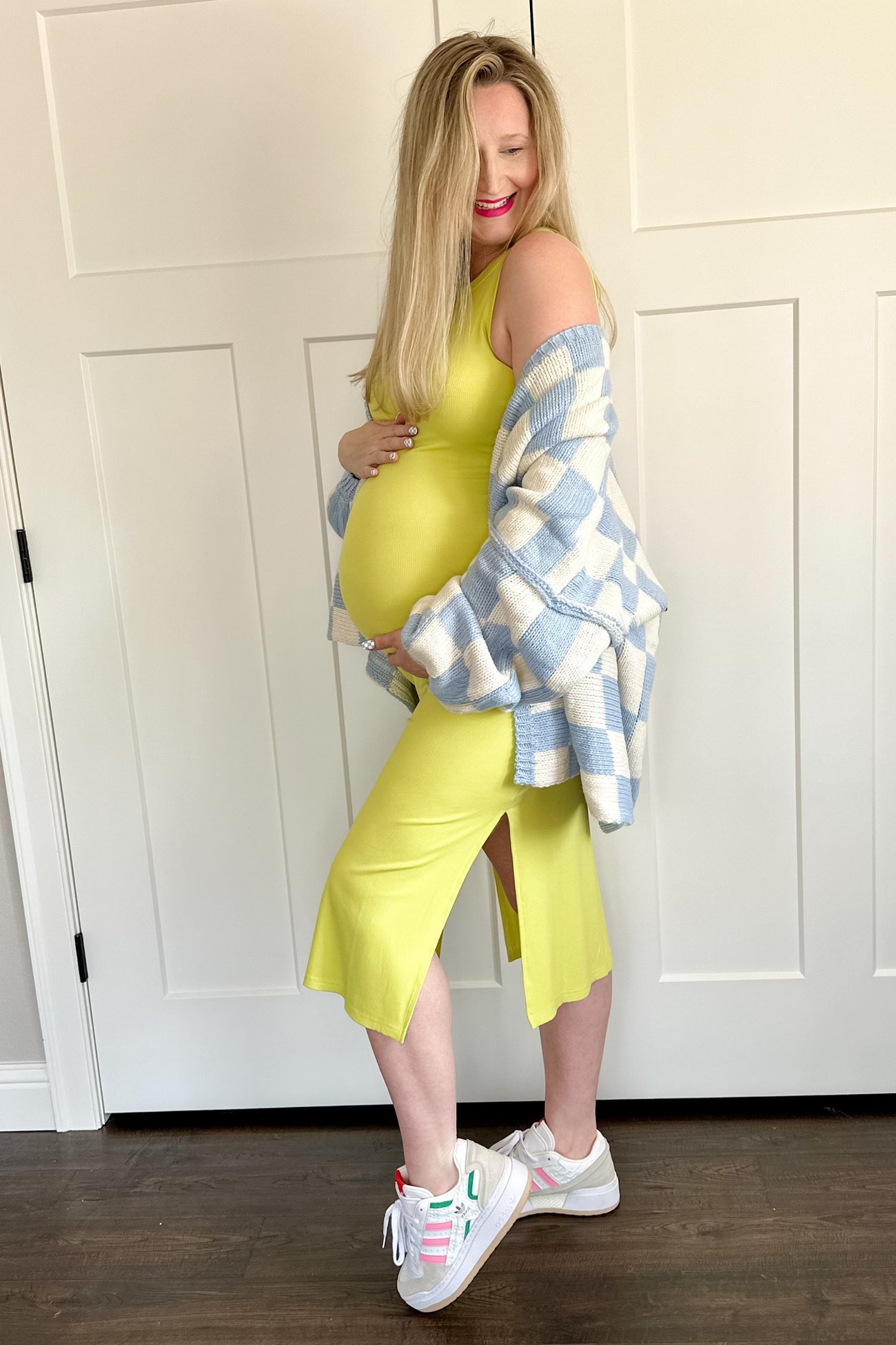 woman wearing a bright yellow maternity dress with a slit, holding her baby bump, and layered with a blue and white checkered cardigan