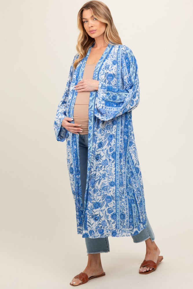 woman wearing a blue floral maternity kimono over a beige top and showing her baby bump while standing