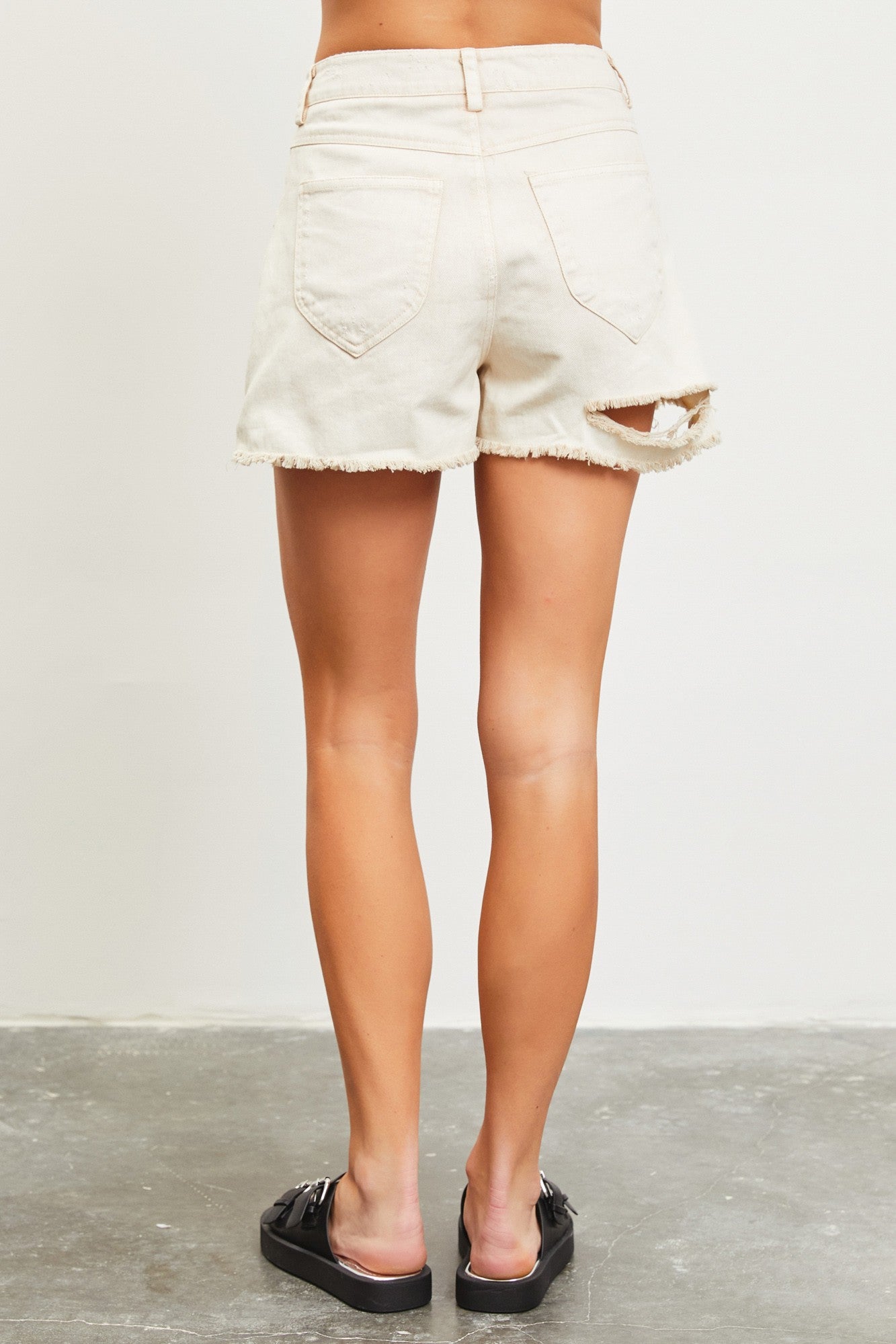 woman wearing light beige distressed denim shorts with frayed hem and cut-out details, viewed from the back