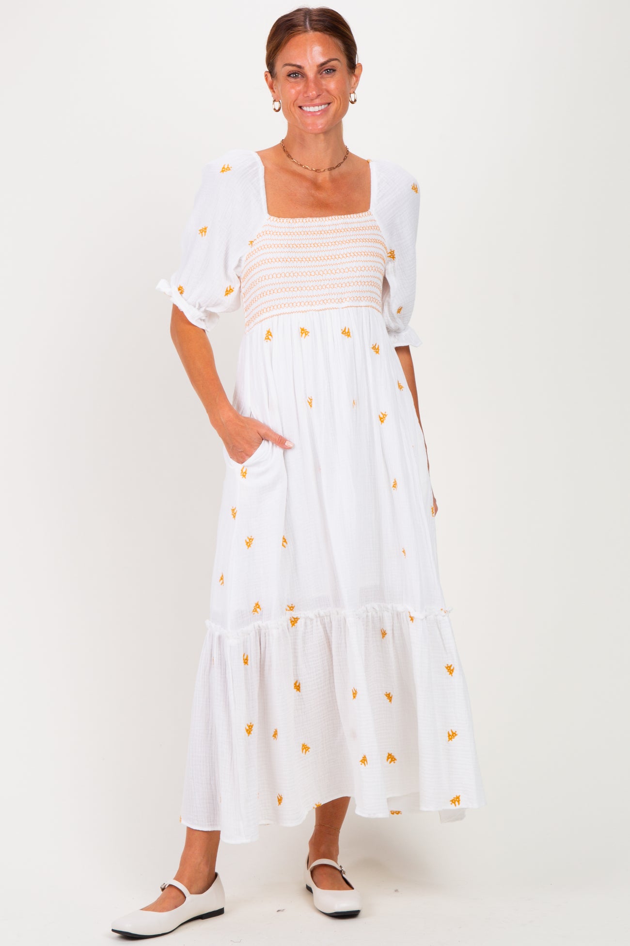 woman wearing a white maxi dress with yellow floral embroidery and puff sleeves, standing with hands in pockets