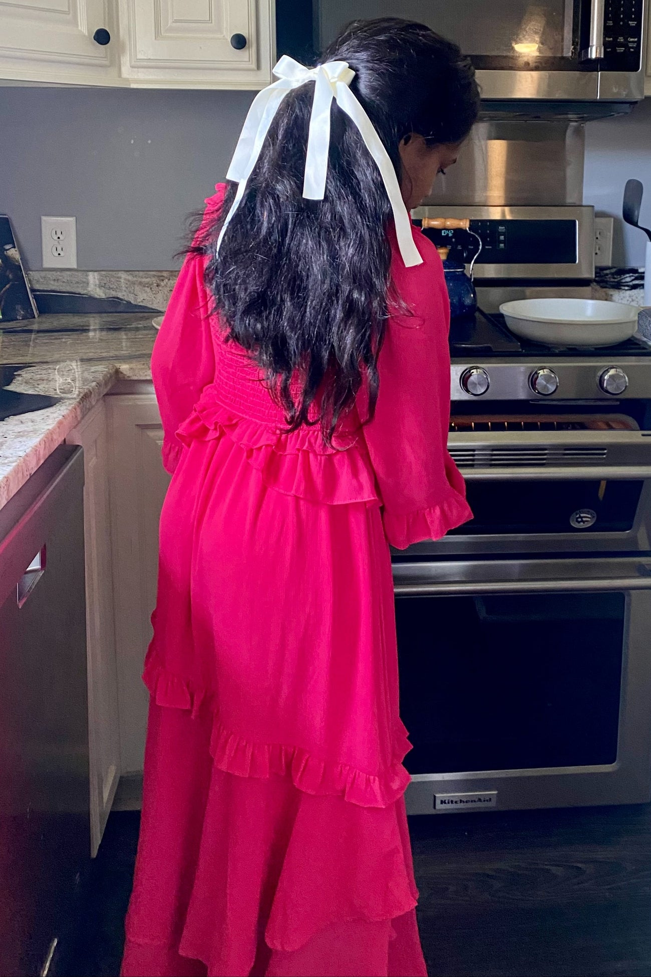 woman wearing a bright pink long sleeve ruffled maxi dress with a white ribbon in her hair, cooking in a kitchen