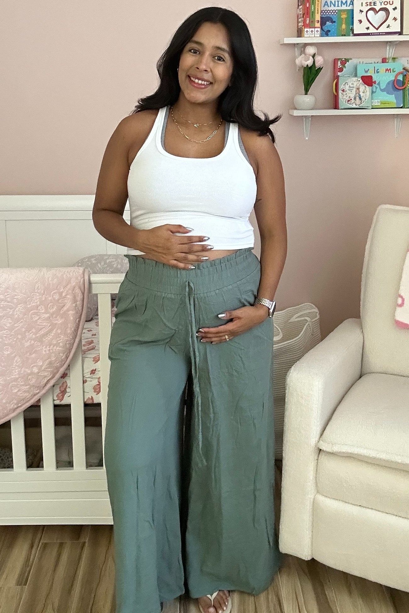 woman wearing a white tank top and green wide-leg maternity pants, holding her baby bump in a nursery setting