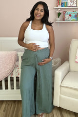woman wearing a white tank top and green wide-leg maternity pants, holding her baby bump in a nursery setting