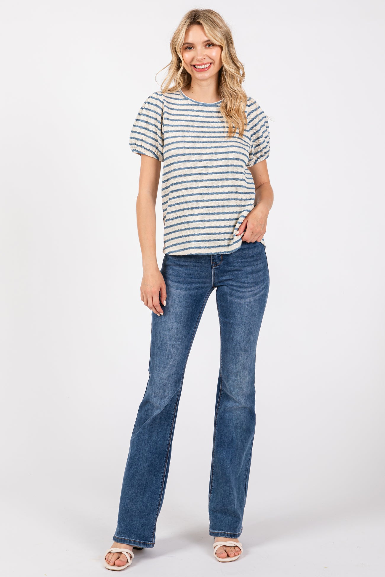 woman wearing a striped short sleeve top and flared jeans, standing and smiling against a white background