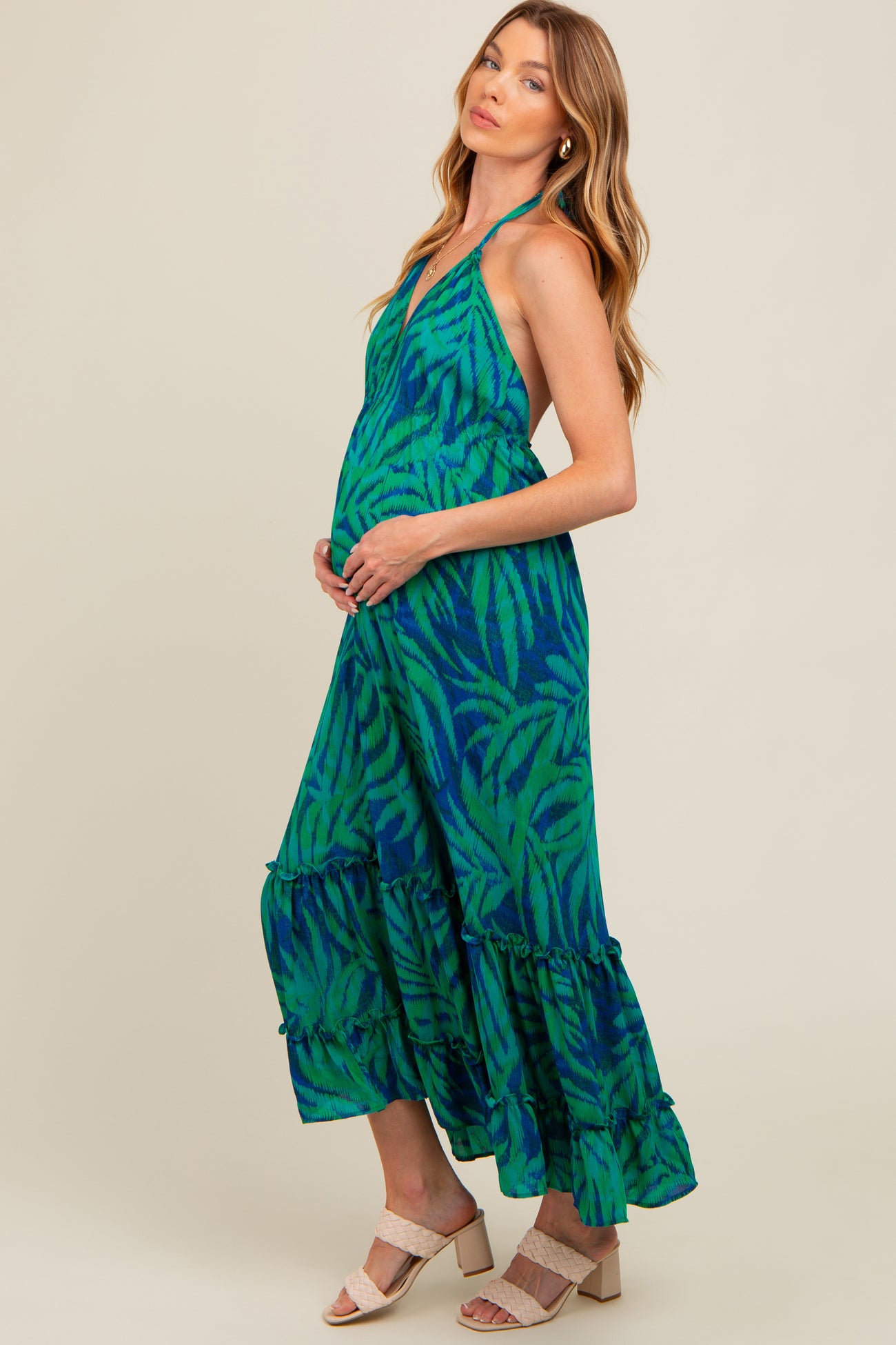 woman wearing a green and blue tropical print halter maternity dress with a ruffled hem and holding her baby bump