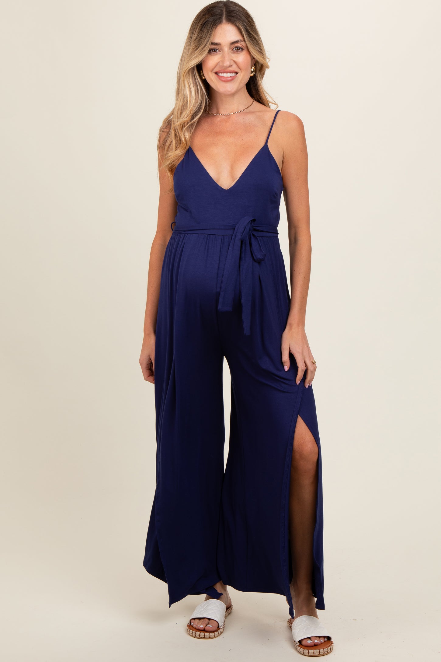 woman wearing a navy blue maternity jumpsuit with a plunging neckline, side slits, and a waist tie, showcasing her baby bump