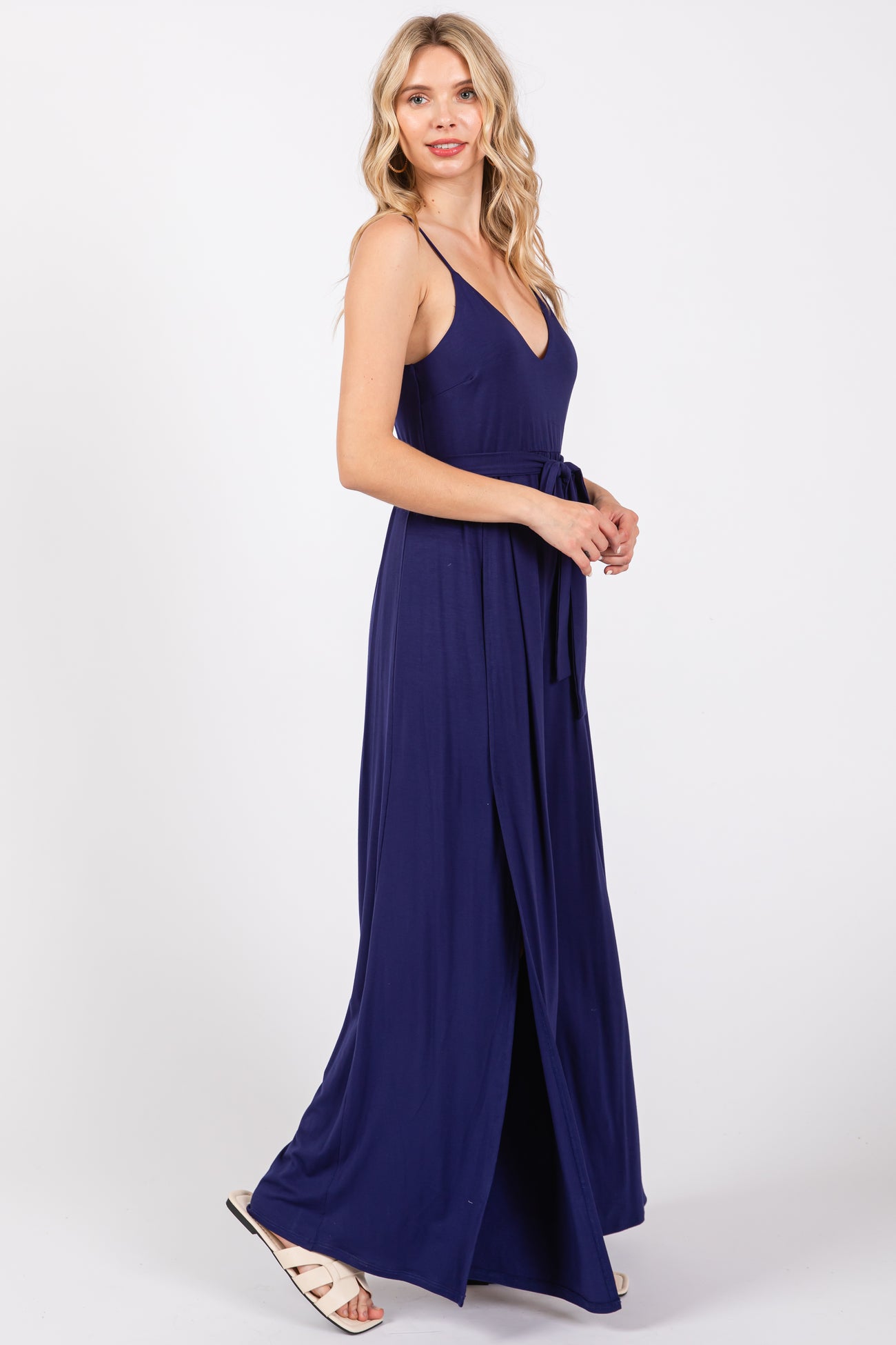 woman wearing a navy blue maxi dress with a deep v-neckline and a tie belt, standing in profile with a slit at the side