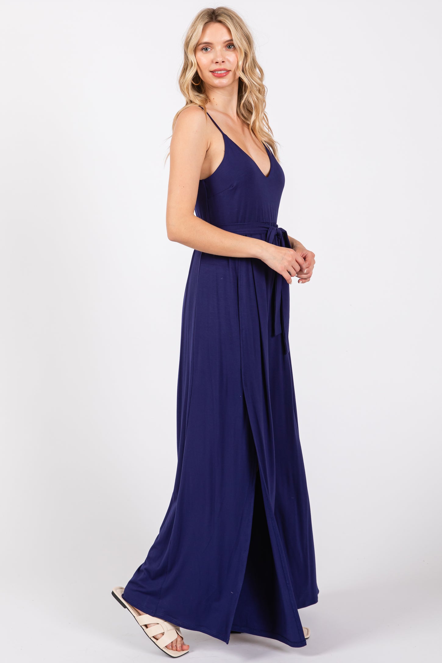 woman wearing a navy blue maxi dress with a deep v-neckline and a tie belt, standing in profile with a slit at the side