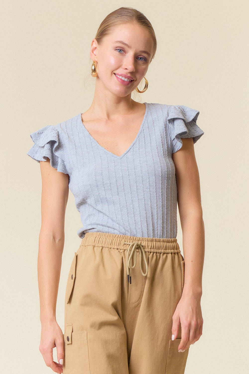 woman wearing a light gray short sleeve top with ruffle details and a v-neckline paired with beige pants