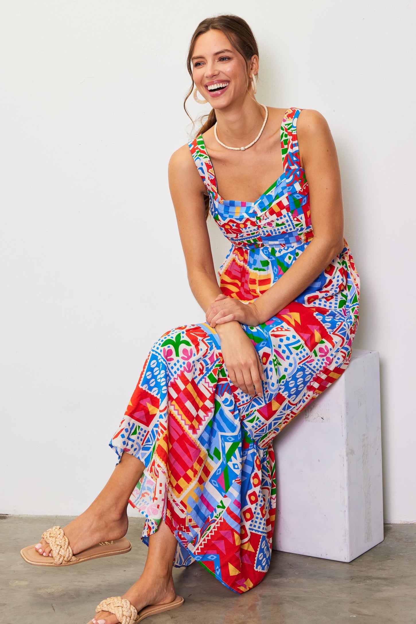 woman wearing a colorful patterned maxi dress with a square neckline and smiling while sitting on a white block