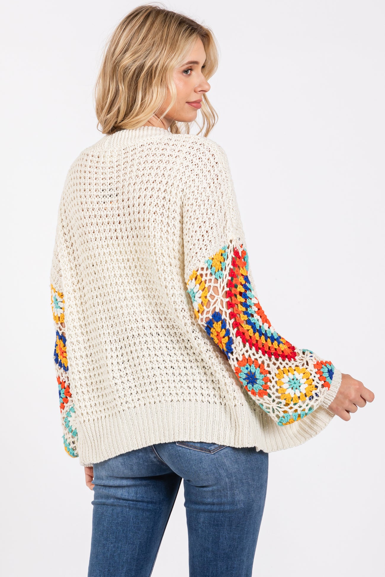 woman wearing a light cream crochet sweater with colorful embroidered sleeves and blue jeans