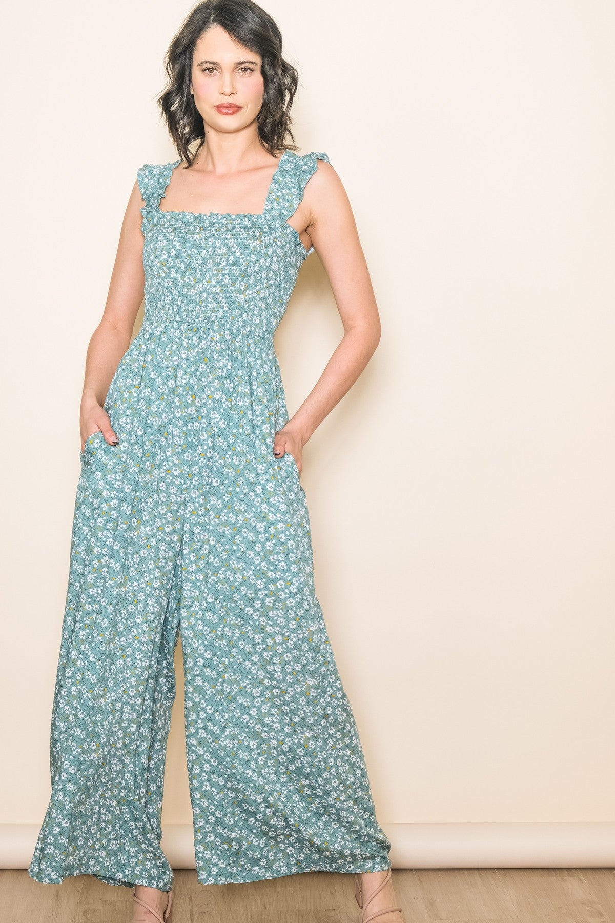 woman wearing a teal floral jumpsuit with square neckline and ruffle straps, posing with hands in pockets