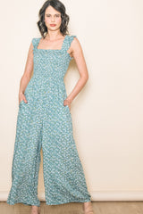 woman wearing a teal floral jumpsuit with square neckline and ruffle straps, posing with hands in pockets