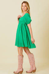 woman wearing a short green puff sleeve dress and pink tall boots, posing sideways with a neutral background