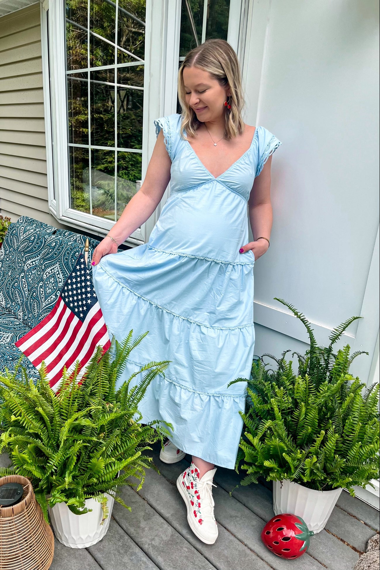 woman wearing a light blue maternity maxi dress with ruffle sleeves and pockets, standing beside potted ferns