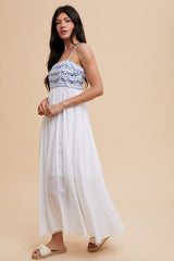 woman wearing a white maxi dress with blue embroidered bodice and spaghetti straps.