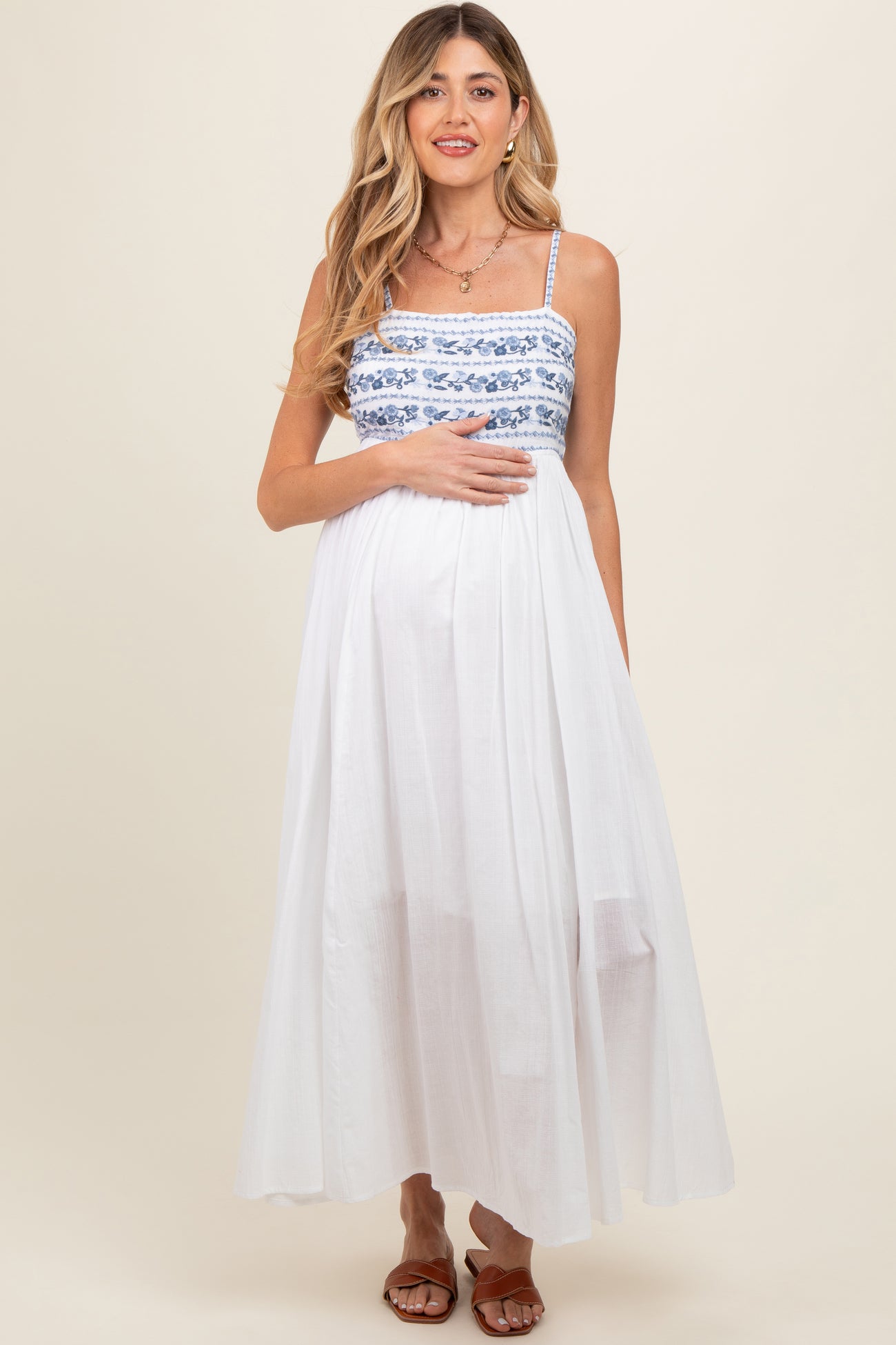 woman wearing a white maxi maternity dress with blue embroidered details and holding her baby bump