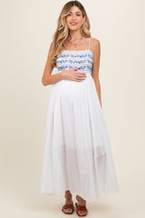 woman wearing a white maxi maternity dress with blue embroidered details and holding her baby bump