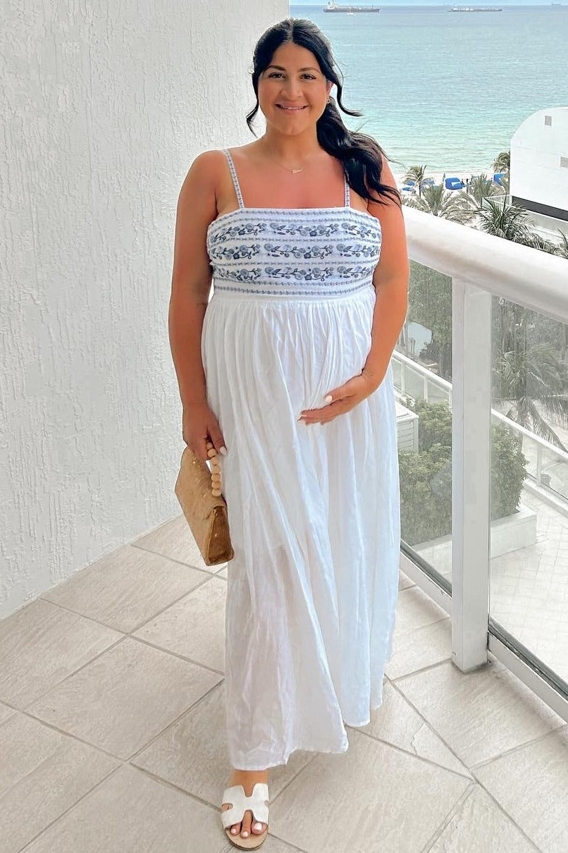 woman wearing a white maternity maxi dress with blue embroidery and holding her baby bump