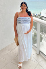 woman wearing a white maternity maxi dress with blue embroidery and holding her baby bump