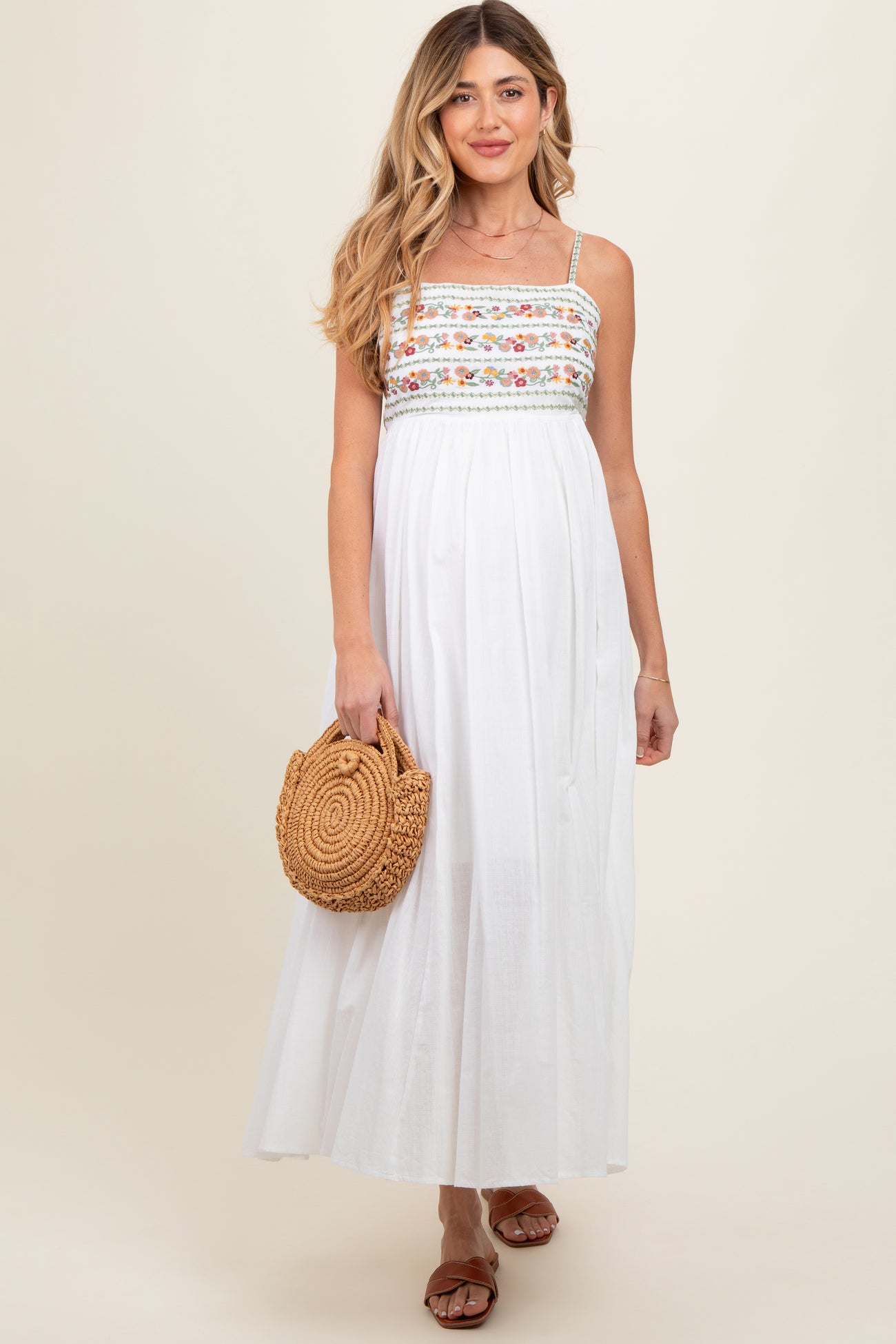 woman wearing a white maternity maxi dress with floral embroidery and holding a straw handbag