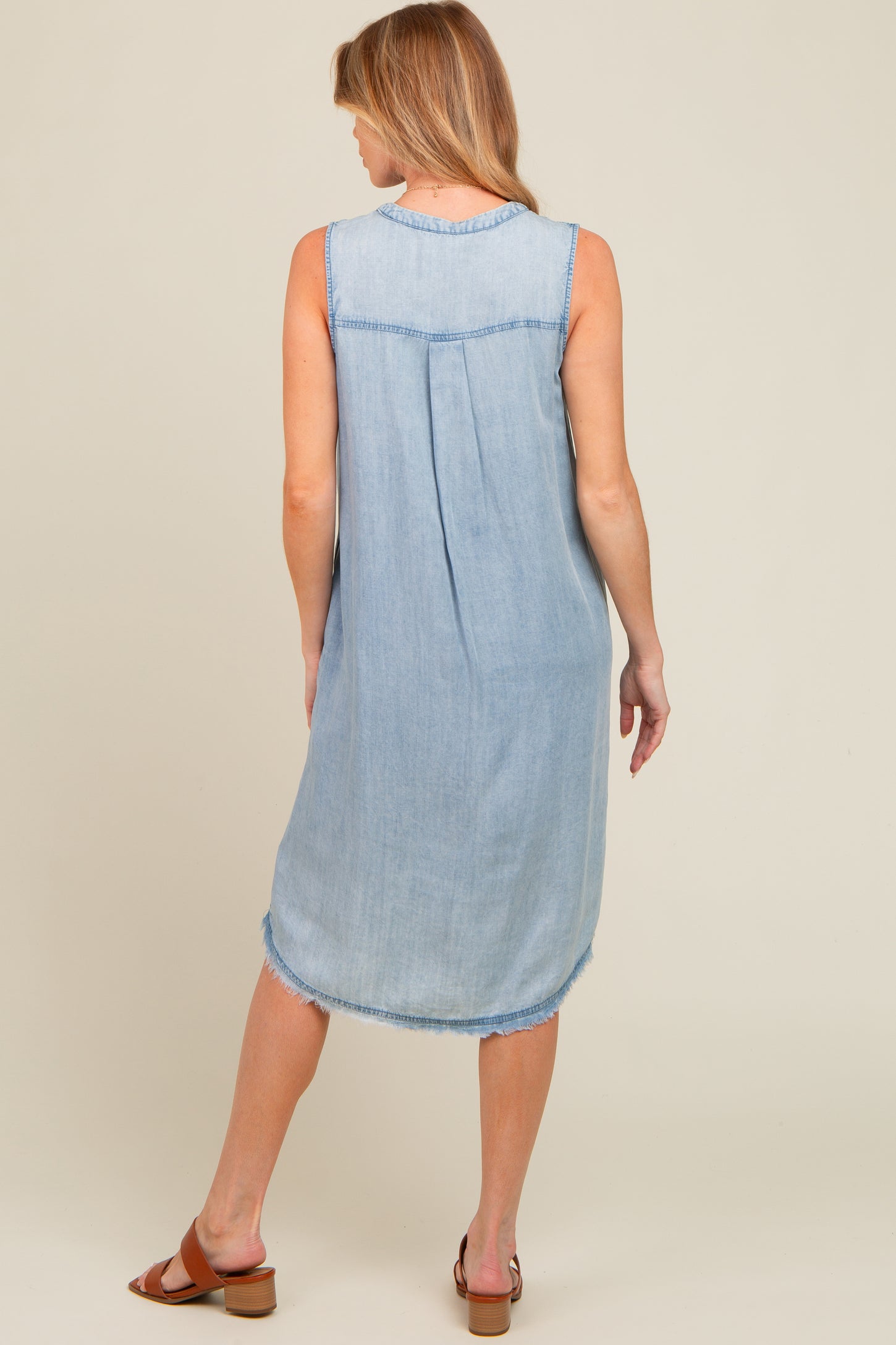 woman wearing a light wash sleeveless denim maternity dress with a relaxed fit and frayed hem, showing the back view