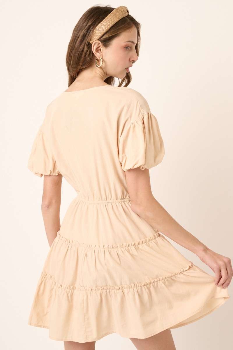 woman wearing a beige puff sleeve tiered mini dress with an elastic waist and ruffled hem