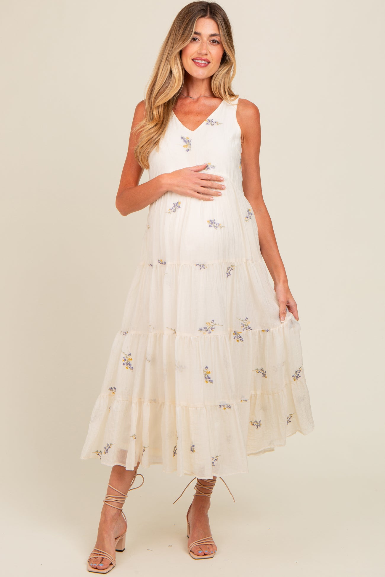 woman wearing a cream maternity dress with floral embroidery and tiered ruffles, holding her baby bump