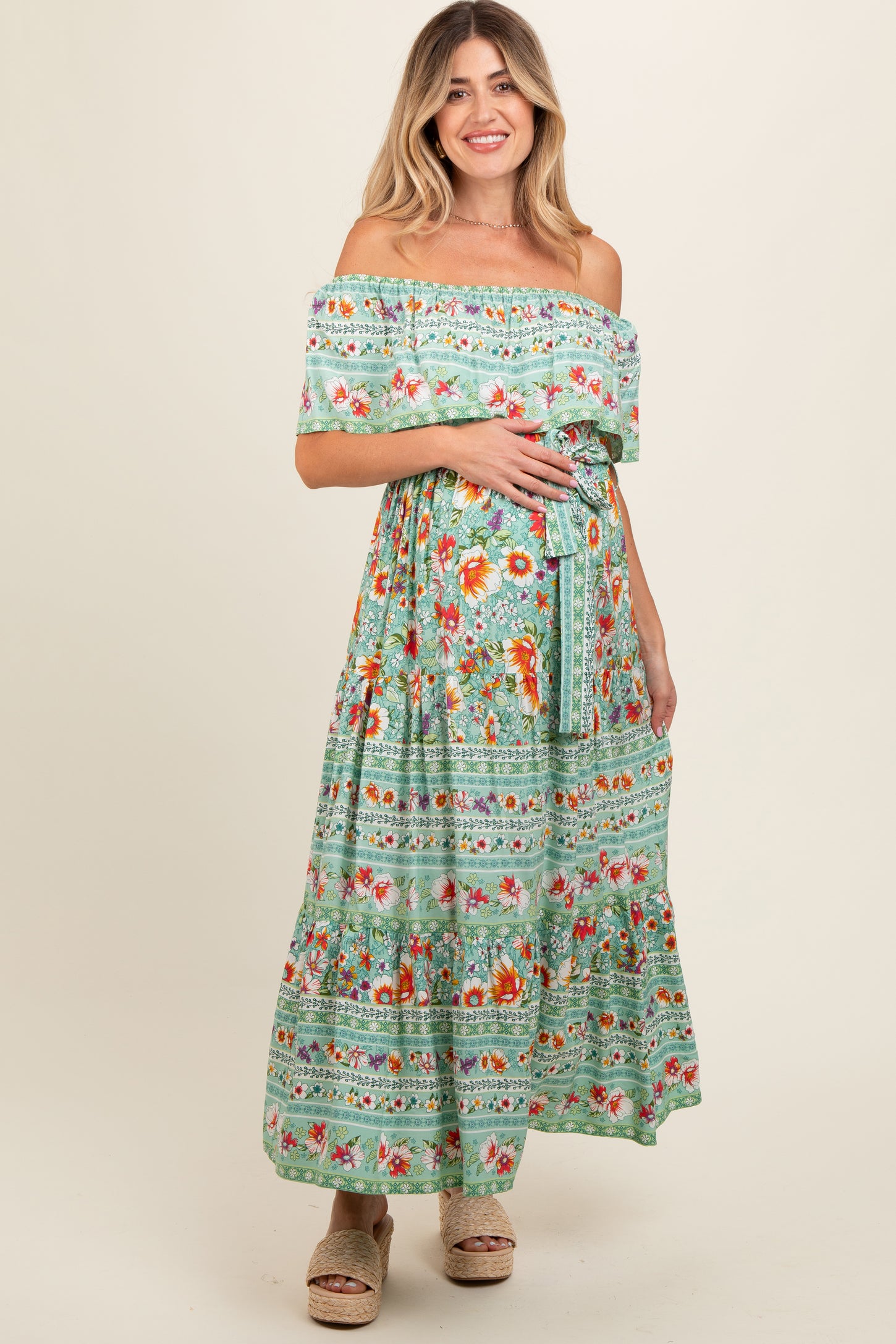 woman wearing a green floral off-the-shoulder maternity maxi dress and holding her baby bump