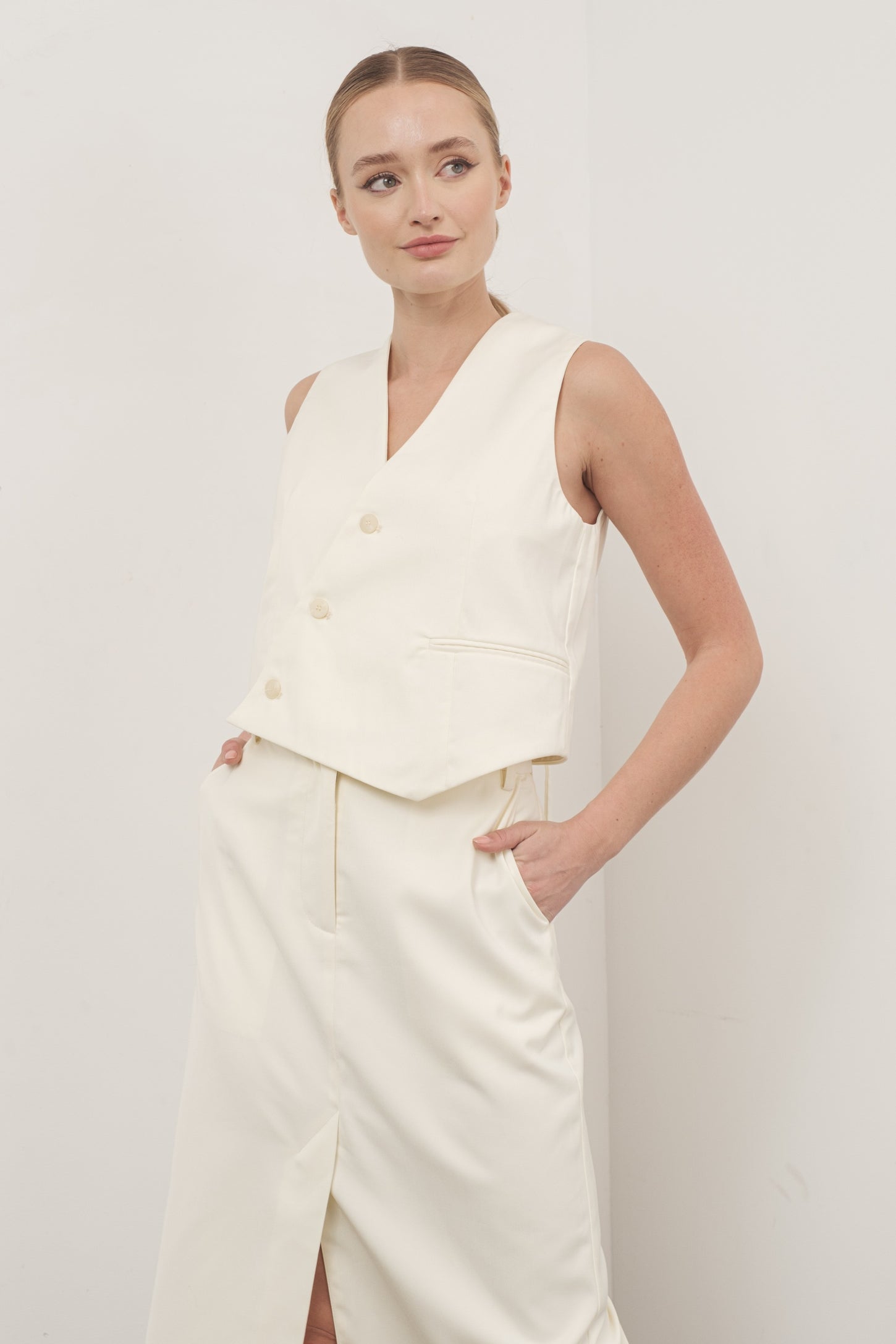 woman wearing a cream sleeveless tailored vest with double buttons and pockets, paired with a matching skirt