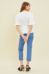 woman wearing a white puff sleeve blouse with a cinched waist and blue cropped jeans, standing with hands behind her back