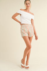 woman wearing a white off-the-shoulder top and colorful patterned shorts, standing with one hand on her hip